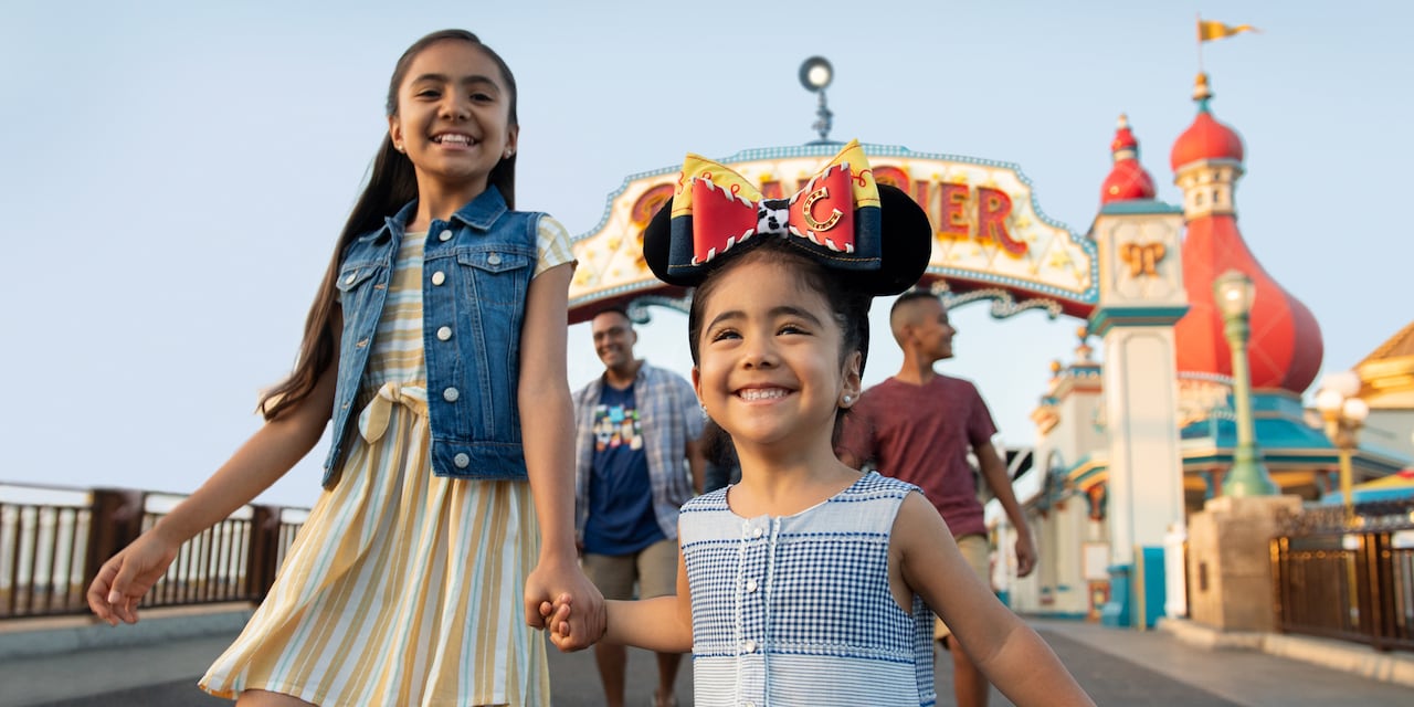 Two smiling girls, the smaller one wearing Micky Ears adorned with a Jesse Bow, hold hands as they exit Paradise Pier ahead of 2 older boys who walk behind them