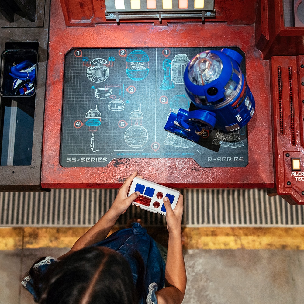Una niña controla su nuevo droide de la serie R sobre una consola de taller con planos de construcción de droides.