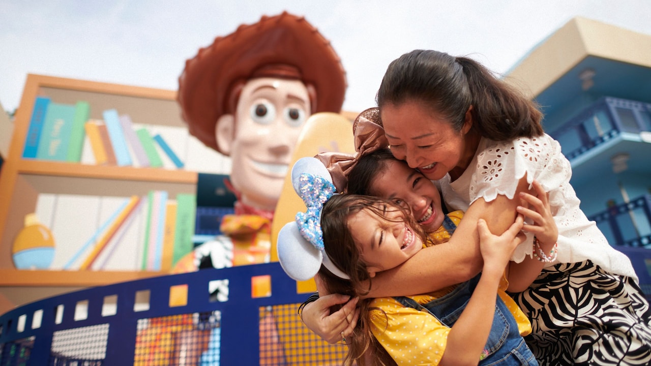 A mother hugging her 2 daughters, each wearing a Minnie Mouse ear headband, near a statue of Sheriff Woody