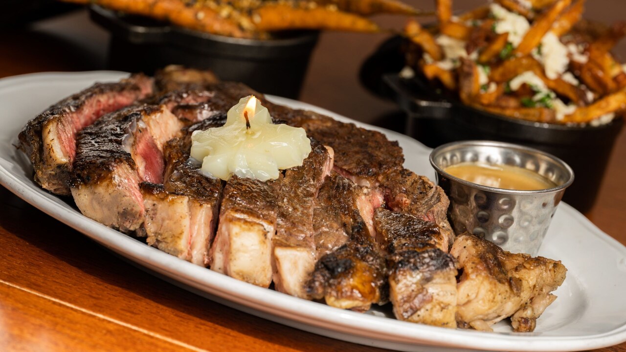 A large steak with a butter pat candle and a ramekin of sauce, served with sides of poutine and roasted carrots