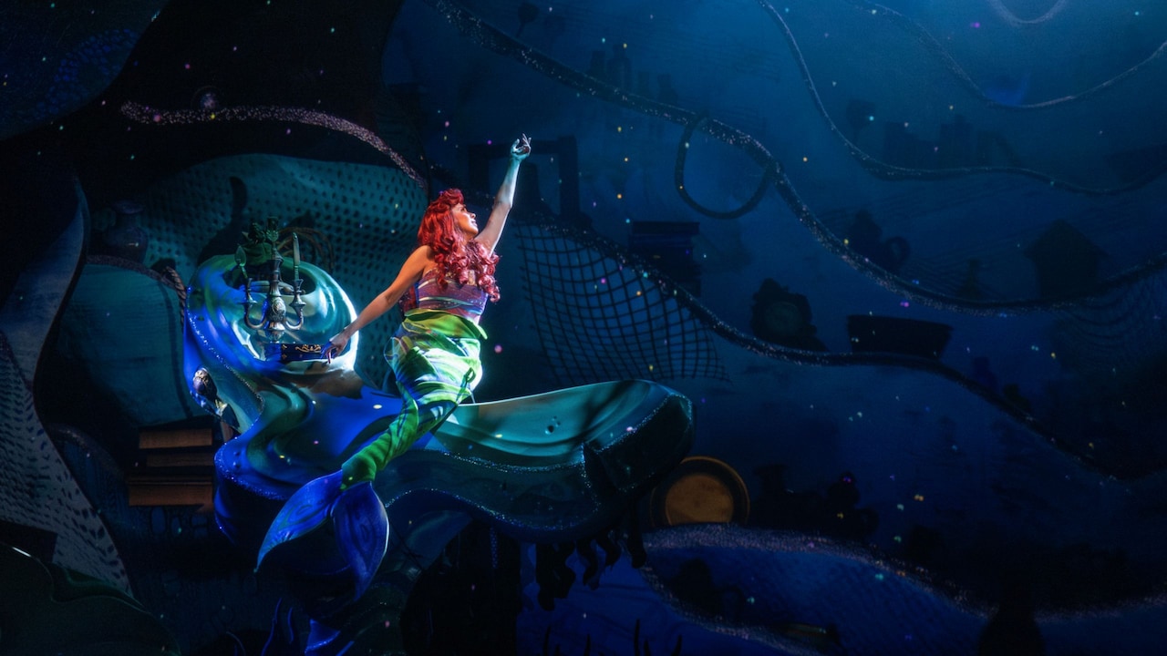 The Little Mermaid sings while perched on the edge of a large rock on the sea floor