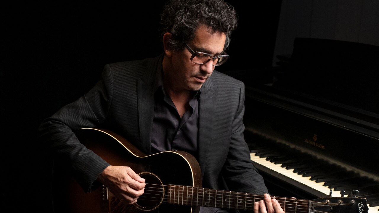 A.J. Croce strumming a guitar while sitting in front of a piano