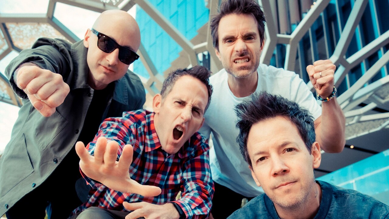 4 members of the group Simple Plan posing while sitting on top of a bench in front of a large abstract sculpture