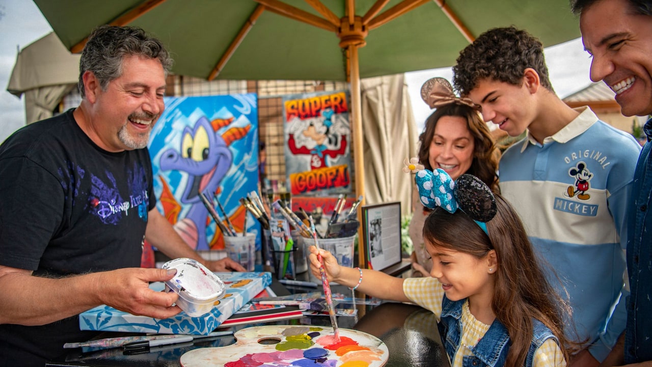 A young girl brushes paint on an artist’s palette, while the artist, the girl’s parents and her brother smile and watch