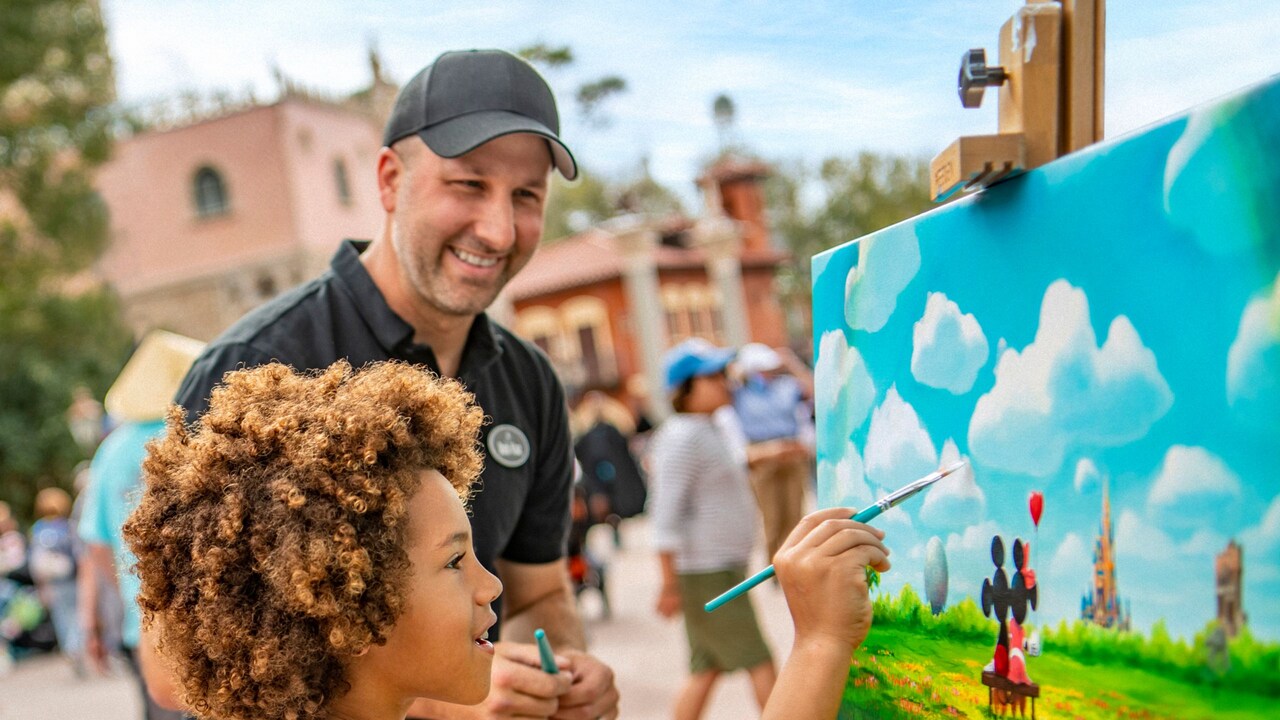 A young boy painting clouds on a painting of Mickey Mouse and Minnie Mouse as an artist looks on at Epcot International Festival of the Arts