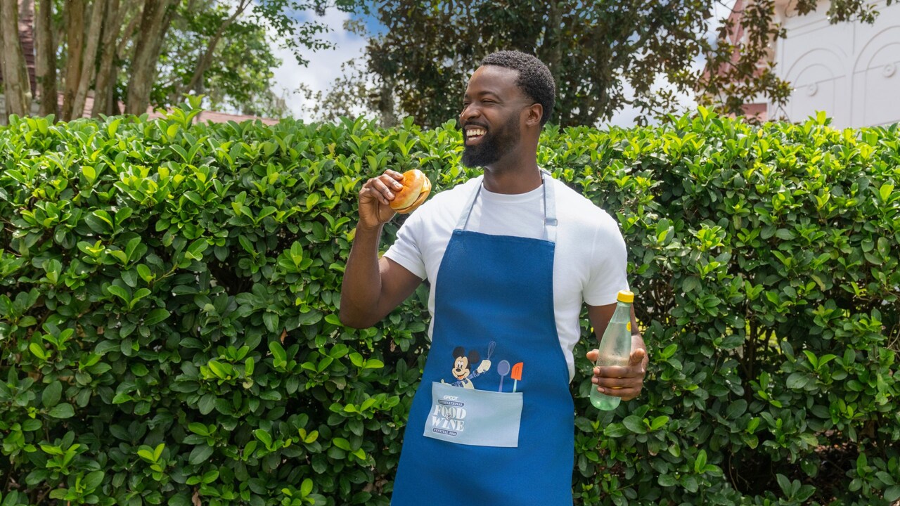 A Guest wearing a tee shirt and an Epcot International Food & Wine Festival apron while holding a burger and a bottle of juice