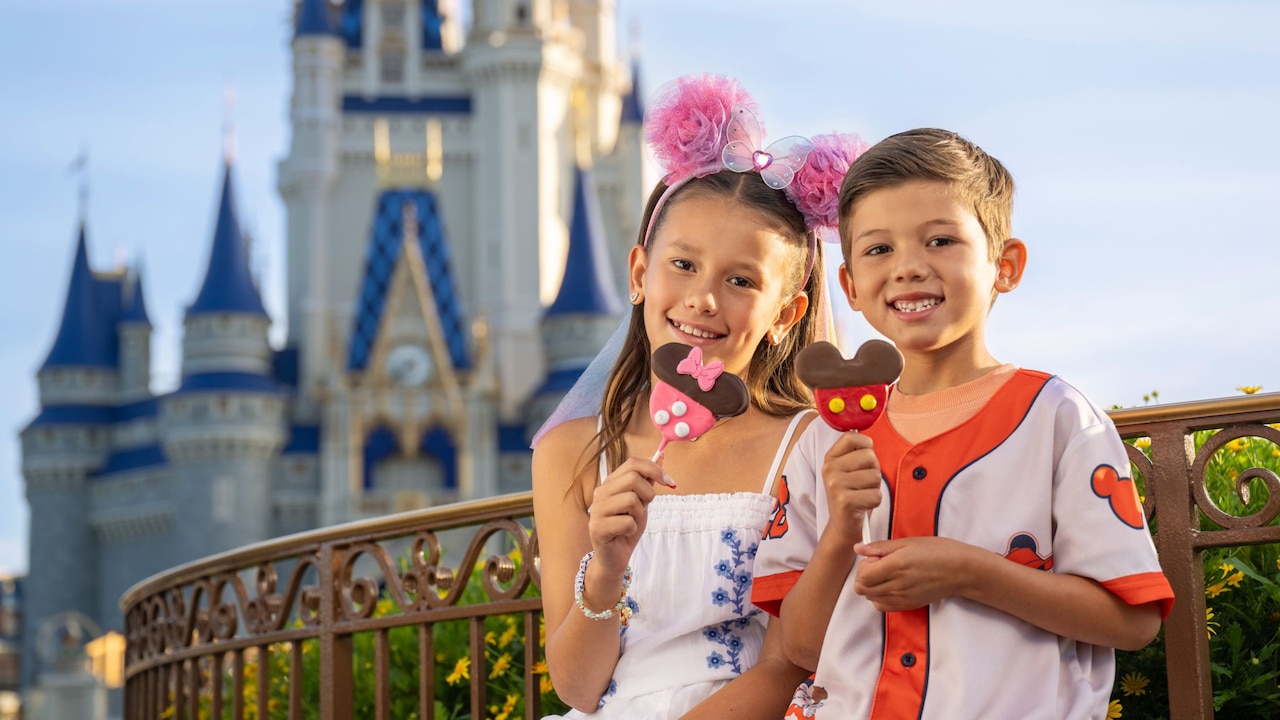 Girl in Minnie Mouse ear headband and boy in Mickey Mouse baseball jersey eating treats by Cinderella Castle