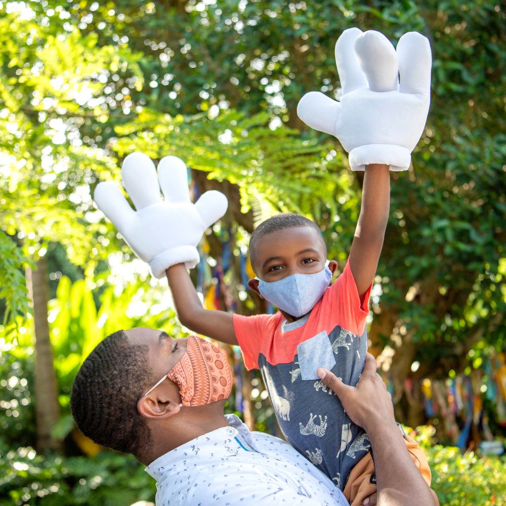 A man holding his son who lifts his arms while wearing oversized Mickey Mouse gloves