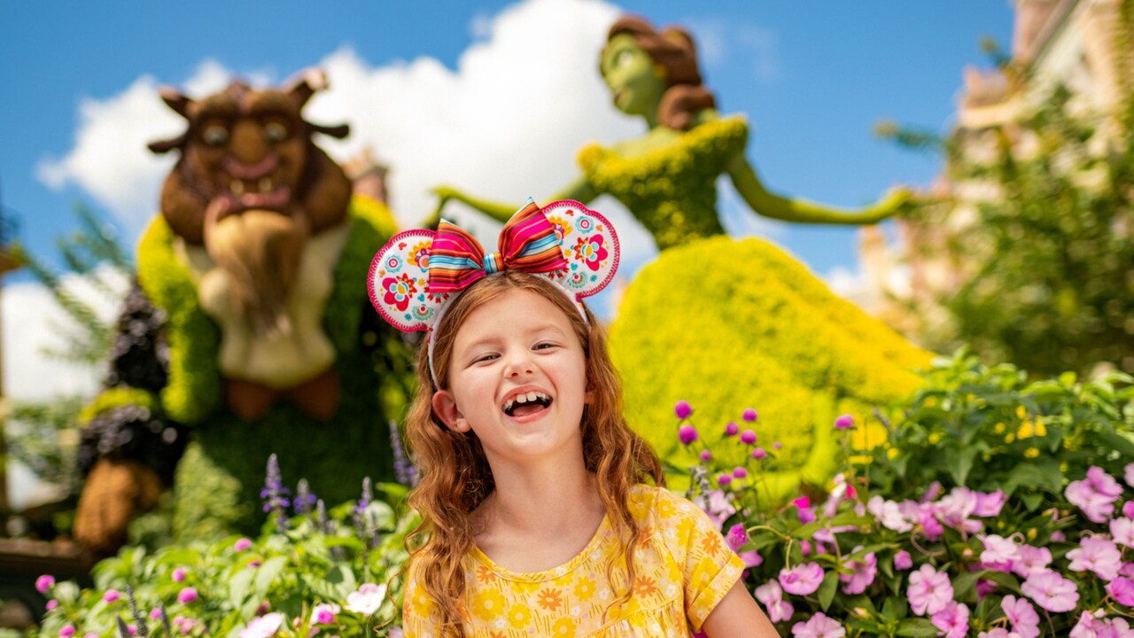 A young girl in a Minnie Mouse ear headband smiles near a Beauty and the Beast topiary in a botanical garden