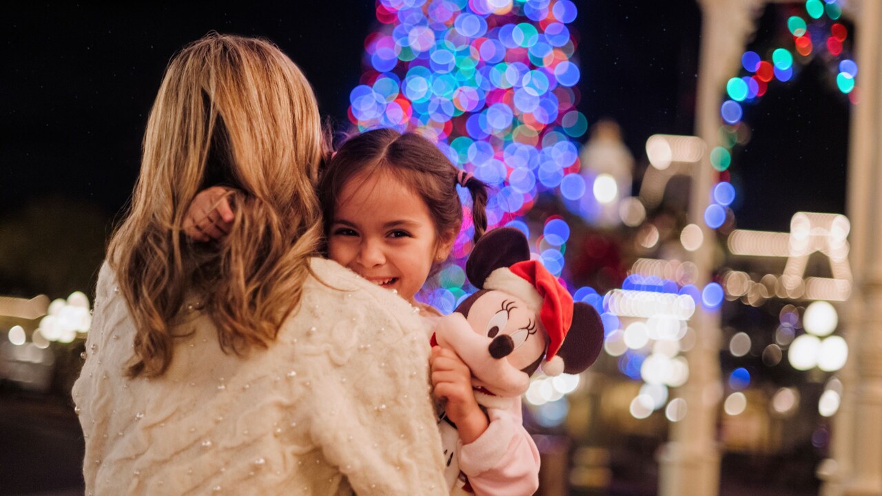 A smiling young girl holds a Minnie Mouse plush toy as she is carried by her mom
