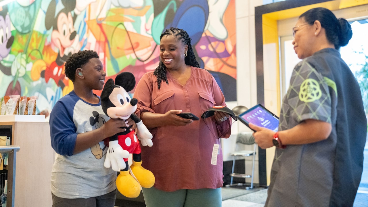 A boy with a hearing aid holds a plush Mickey Mouse, smiling with a woman and female Cast Member.