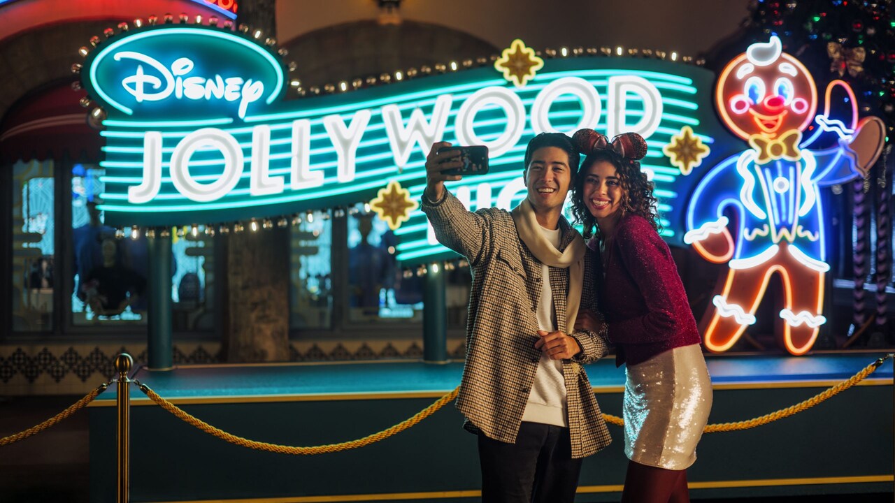 A couple taking a selfie in front of an illuminated sign that says, 'Disney Jollywood Nights'