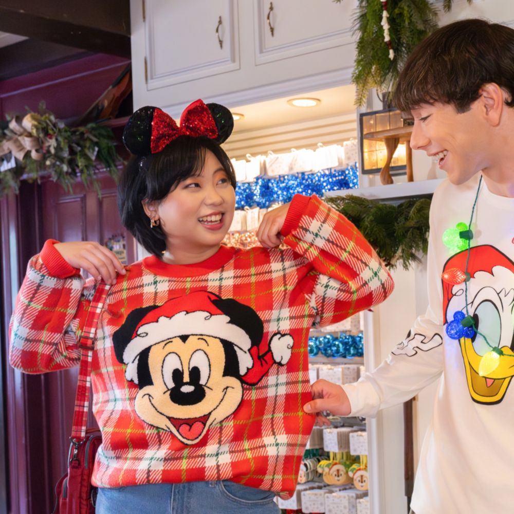 Two Guests wearing Christmas apparel including a sweater featuring Mickey Mouse, a long sleeve shirt featuring Donald Duck, Pluto and Goofy, a Minnie Mouse ear headband and a string light necklace
