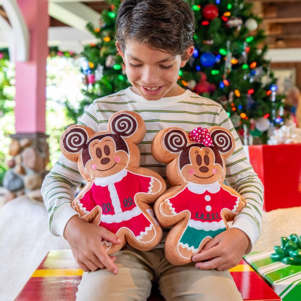 A smiling boy holding gingerbread shaped Mickey Mouse and Minnie Mouse plush pillows near a Christmas tree and wrapped presents