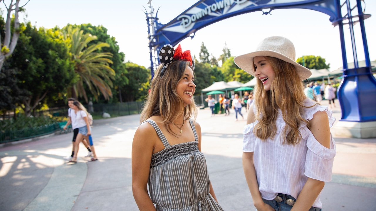 Two young women standing in front of the Downtown Disney arch at the Disneyland Resort share a laugh