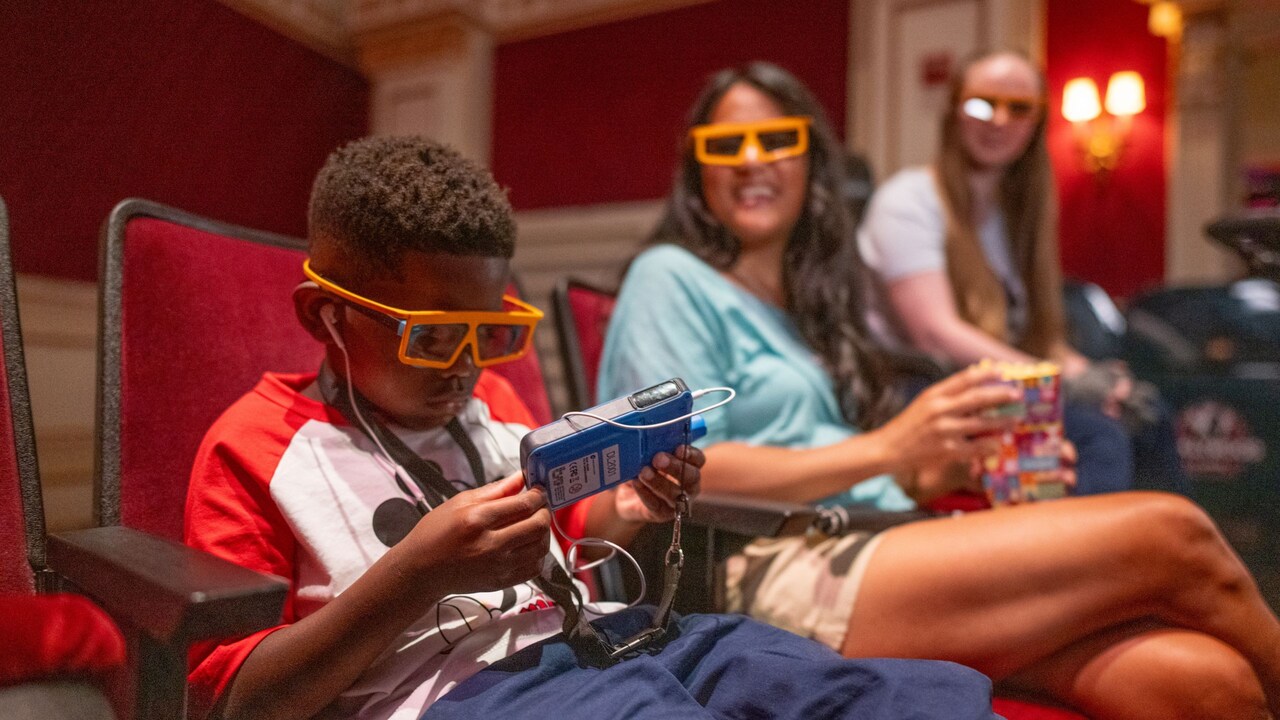 Un niño con gafas 3D se sienta en un cine mientras sostiene y observa el dispositivo portátil Disney.