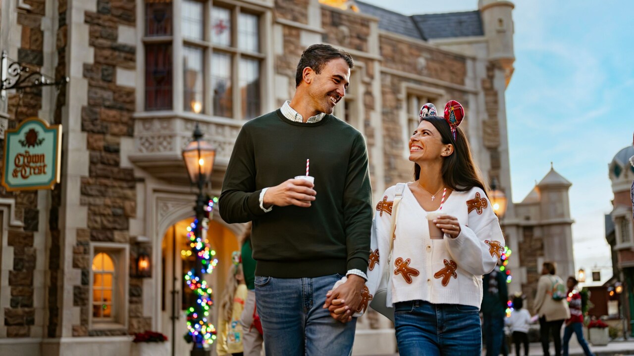 A couple with drinks holds hands as they stroll past lampposts decorated with twinkling holiday lights