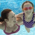 A smiling mother has an arm around her daughter’s waist as they swim underwater in a pool