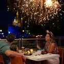 A family of 4 dine on an outdoor terrace as fireworks burst above Cinderella Castle in the background