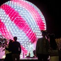 Epcot Guests look up at Spaceship Earth, which is lit up with pink and white diagonal stripes