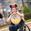 Una joven sonriente, sentada en una silla de ruedas, lleva una diadema con orejas de Minnie y sostiene un artículo conmemorativo con una imagen de Fab Five y las palabras, “Primera visita de Walt Disney World”