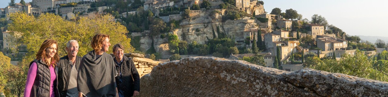 A group of 4 people walk along a stone wall with the mountainside village of Les Baux-de-Provence, France in the background