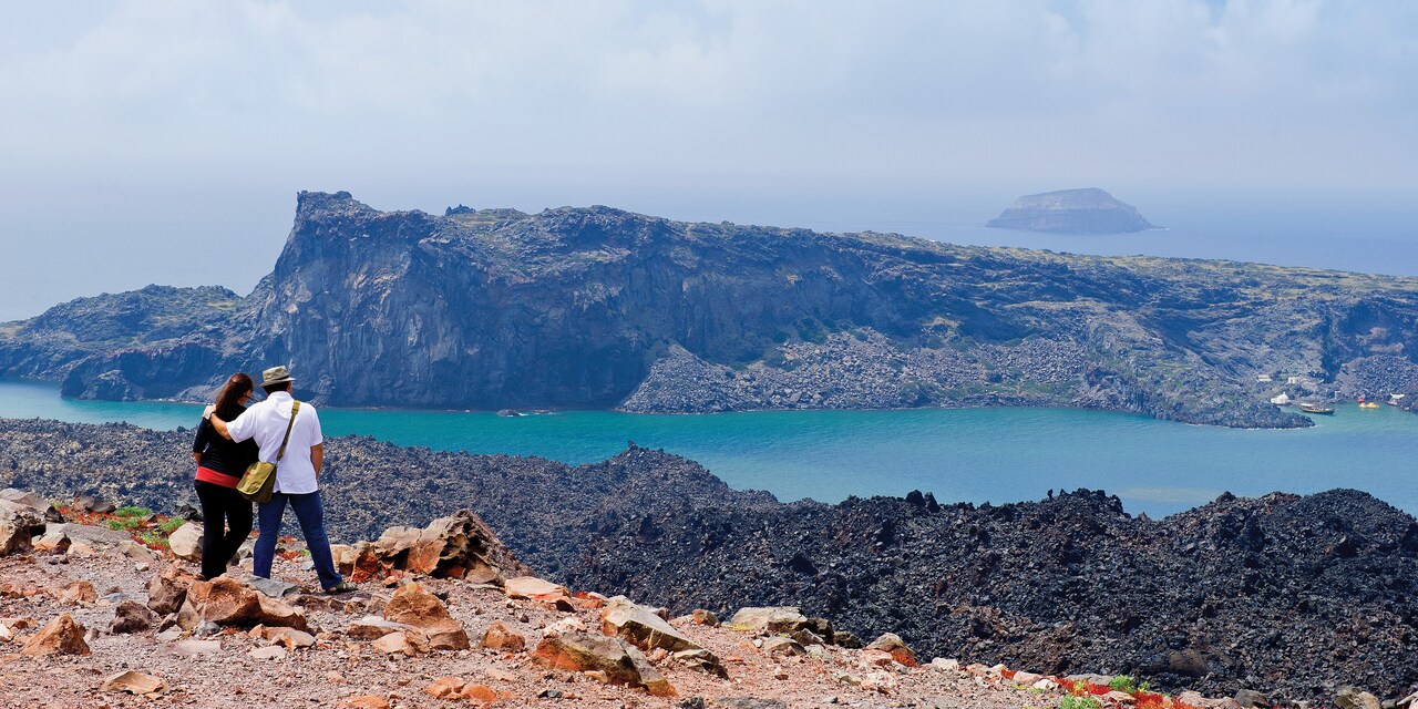 A couple studies a small volcanic island separated from them by a narrow waterway A couple studies a small volcanic island separated from them by a narrow waterway