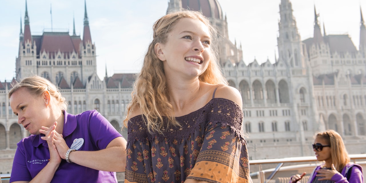 A smiling teenage girl stands near an Adventure Guide with a name tag that says “Stephanie” on the deck of a river boat as it sails past the Gothic Hungarian Parliament Building