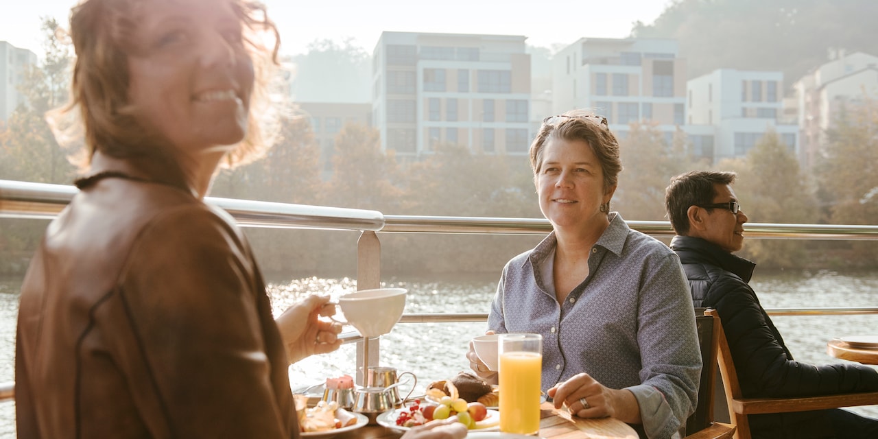 A woman holding a coffee cup looks over her shoulder during a meal with another woman at a table on the top deck of a river boat as it sails past buildings on the nearby shore