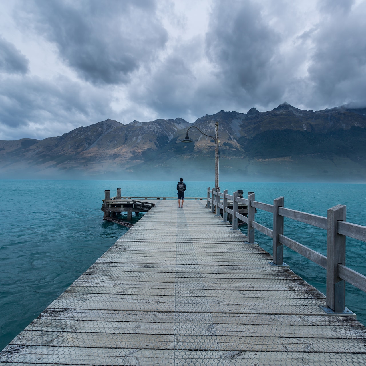 A person stands at the end of a long wooden dock that leads into the sea near a mountain range