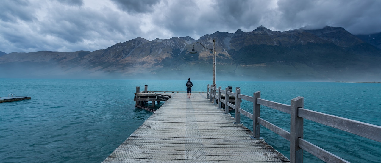 A person stands at the end of a long wooden dock that leads into the sea near a mountain range