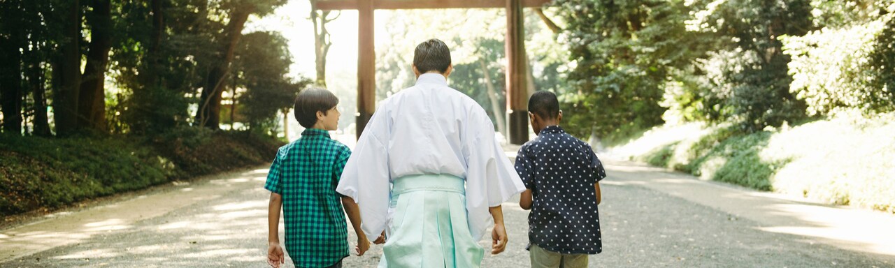 Two boys and a monk walk down a path towards a tojii gate at Meiji Shrine 
