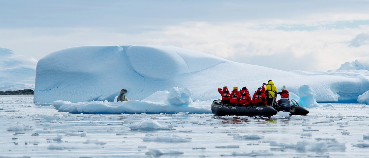 Seven people clad in parkas on a Zodiac boat take pictures of a seal on an iceberg