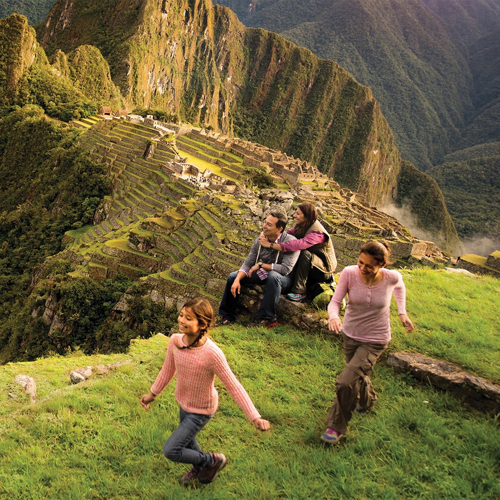 A family of 4 on a grassy area in a mountain range overlooking the ruins of Machu Picchu in Peru