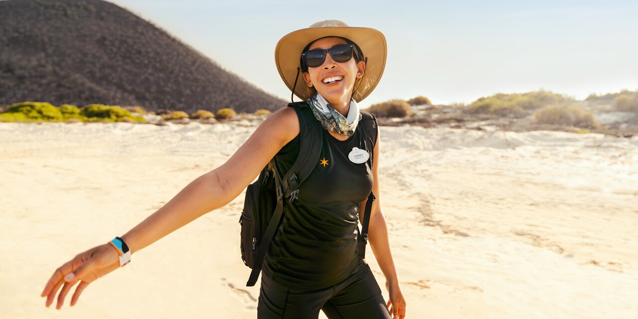 A woman wearing a floppy hat, sunglasses and an Adventures by Disney nametag stands on the sand