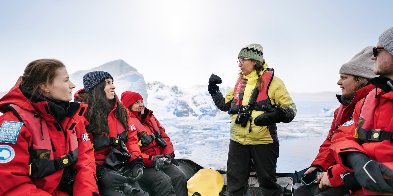 A woman in a knit beanie stands while addressing a group of Adventurers on a boat near icebergs