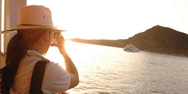 A woman takes a picture of a boat on water with a hill behind it A woman takes a picture of a boat on water with a hill behind it