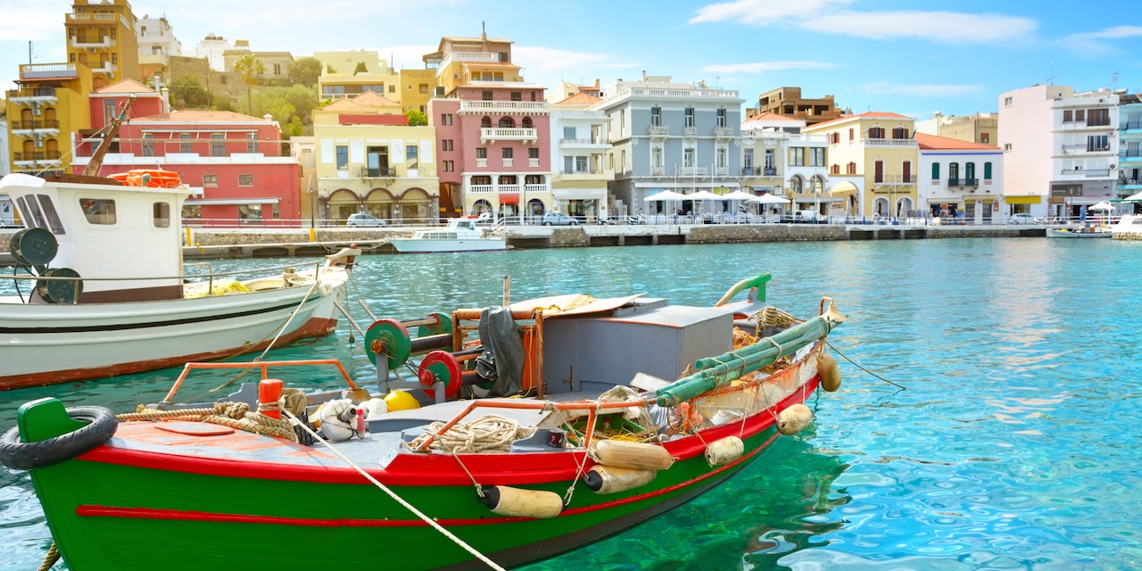 Two boats anchored in a harbor near a street lined with colorful houses in Crete