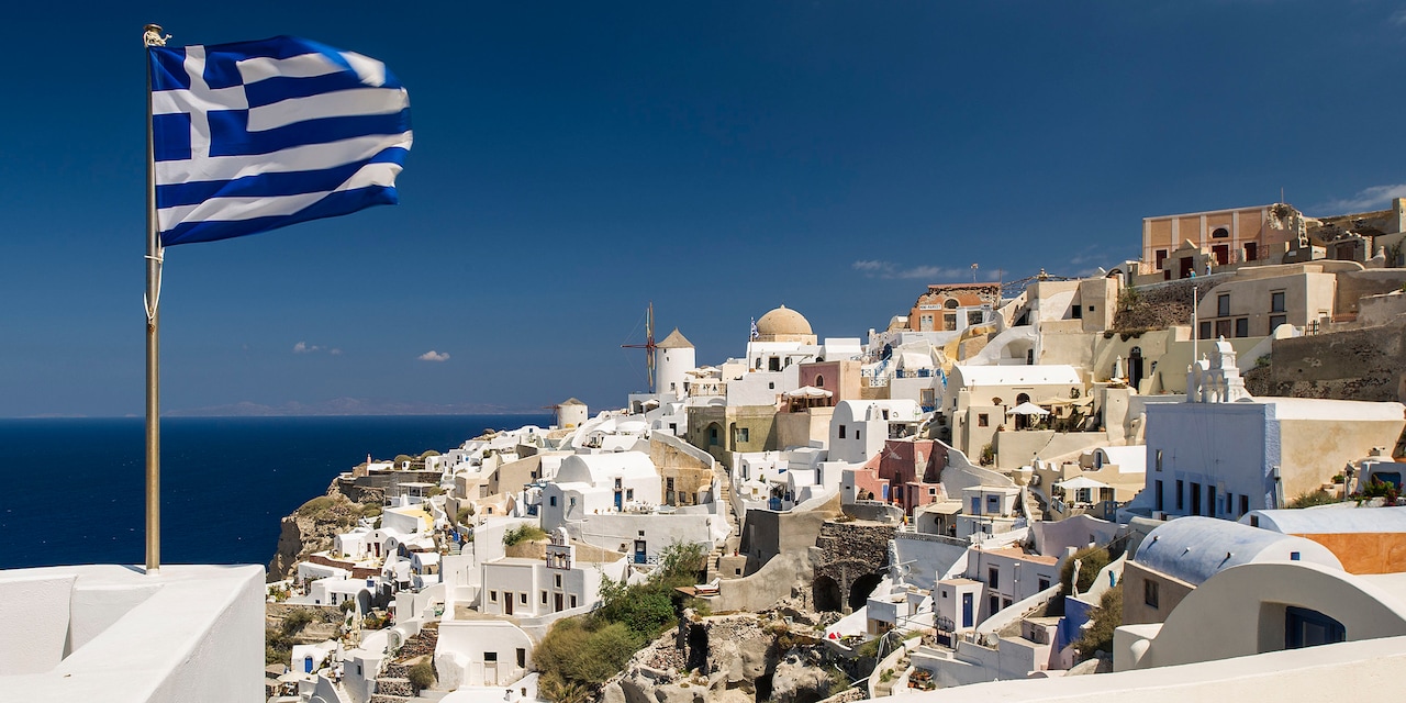 The Greek flag waves from a balcony with the city of Crete and the Aegean Sea in the background