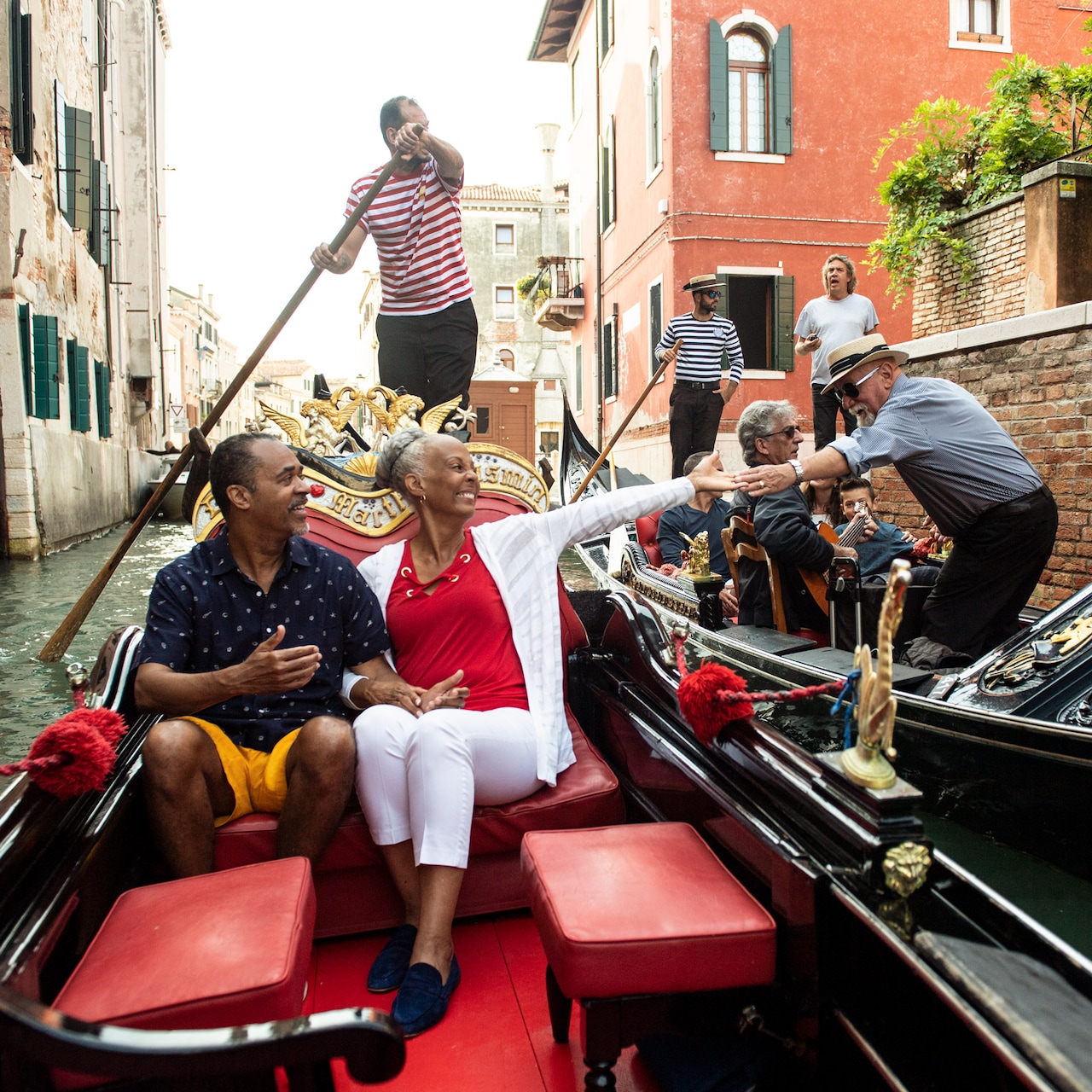 A gondolier rows a couple in a gondola who greet a man in a nearby boat on a canal in Venice, Italy