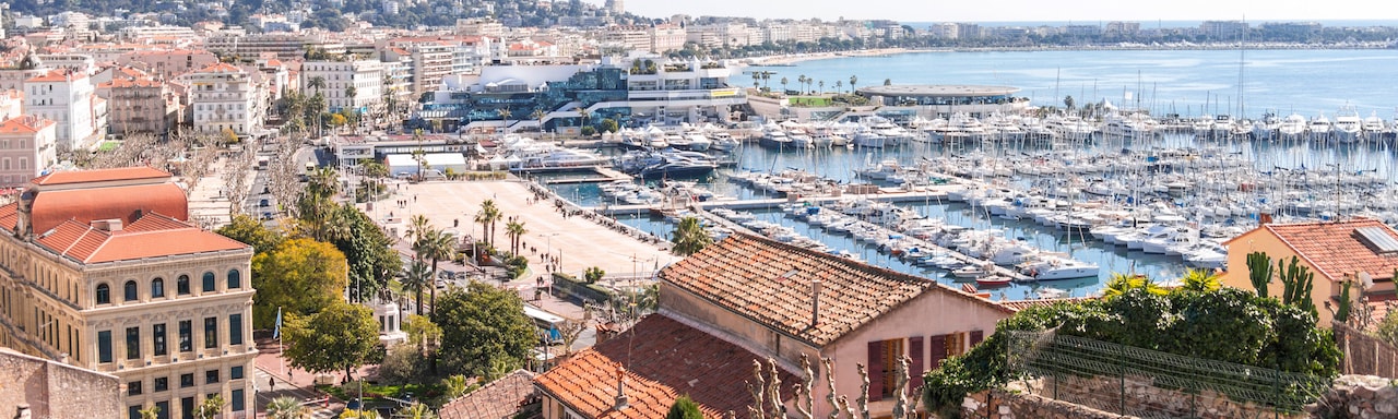 Boats docked in the Cannes Harbor with multi-storied, tile roof buildings hugging the shore Boats docked in the Cannes Harbor with multi-storied, tile roof buildings hugging the shore