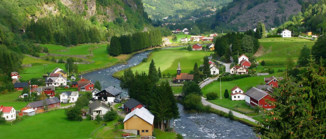 Farmhouses and a church on either side of a creek in a pastoral valley surrounded by mountains 