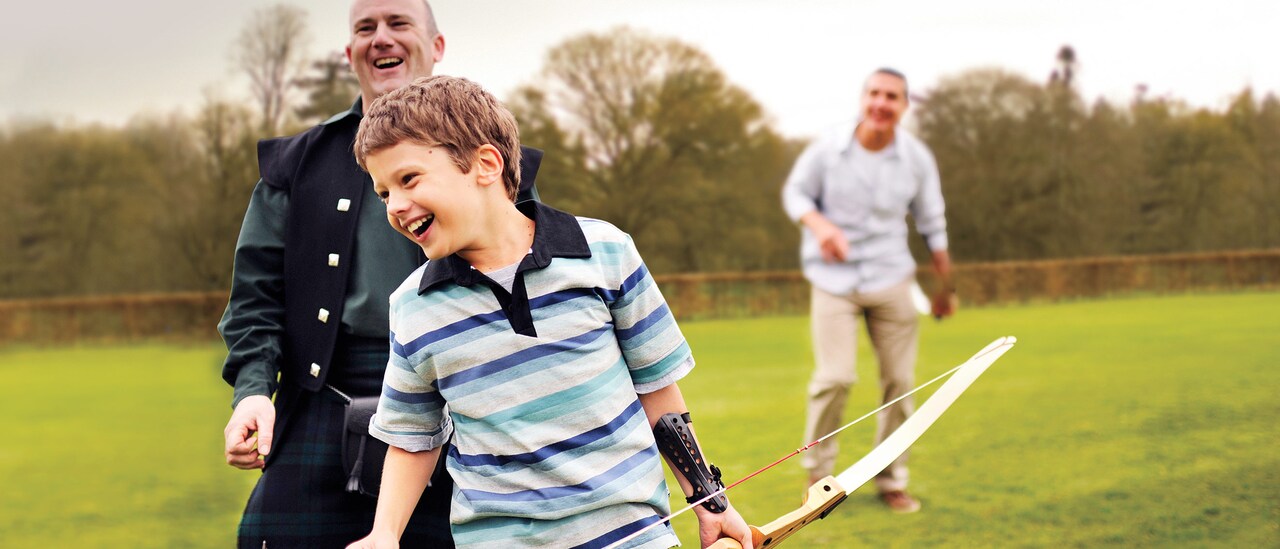 A little boy tries his hand at archery while a teacher and his father watch
