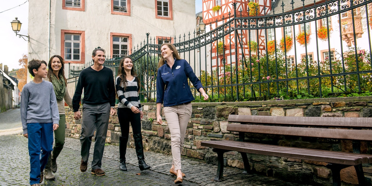 An Adventure Guide and a family of 4 pass a wooden bench and an iron fence as they walk on a cobblestone street An Adventure Guide and a family of 4 pass a wooden bench and an iron fence as they walk on a cobblestone street