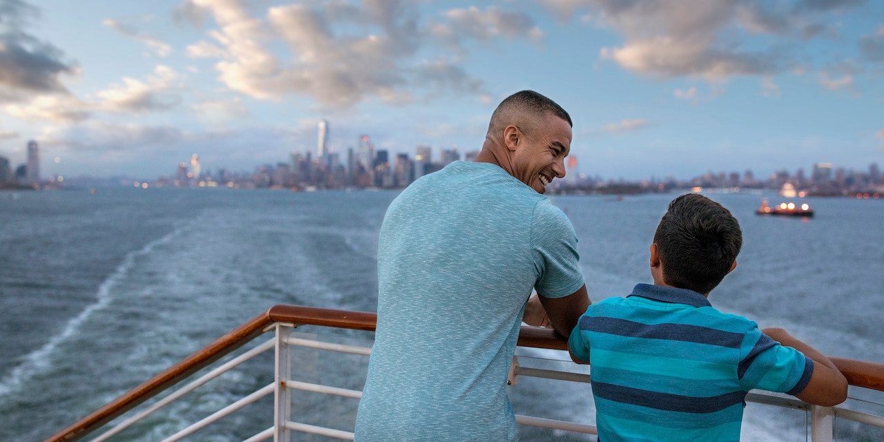 A father and his son look at a distant city from the railing of a ship sailing over a body of water A father and his son look at a distant city from the railing of a ship sailing over a body of water