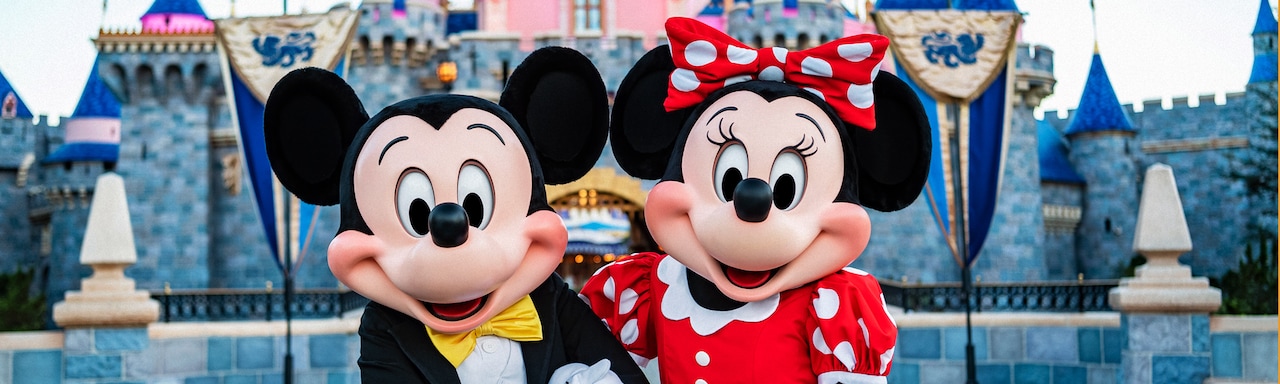 Mickey Mouse and Minnie Mouse stand arm in arm in front of Sleeping Beauty Castle at Disneyland Park