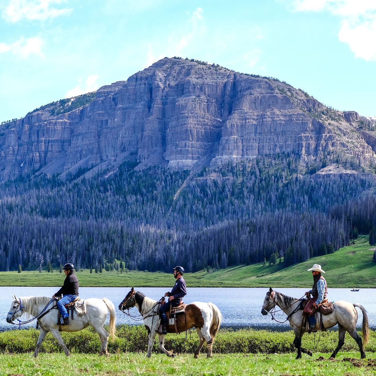 Three people on horseback ride across a river from a mountain with a tree-lined base Three people on horseback ride across a river from a mountain with a tree-lined base