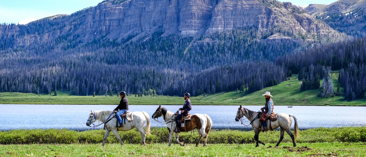Three people on horseback ride across a river from a mountain with a tree-lined base