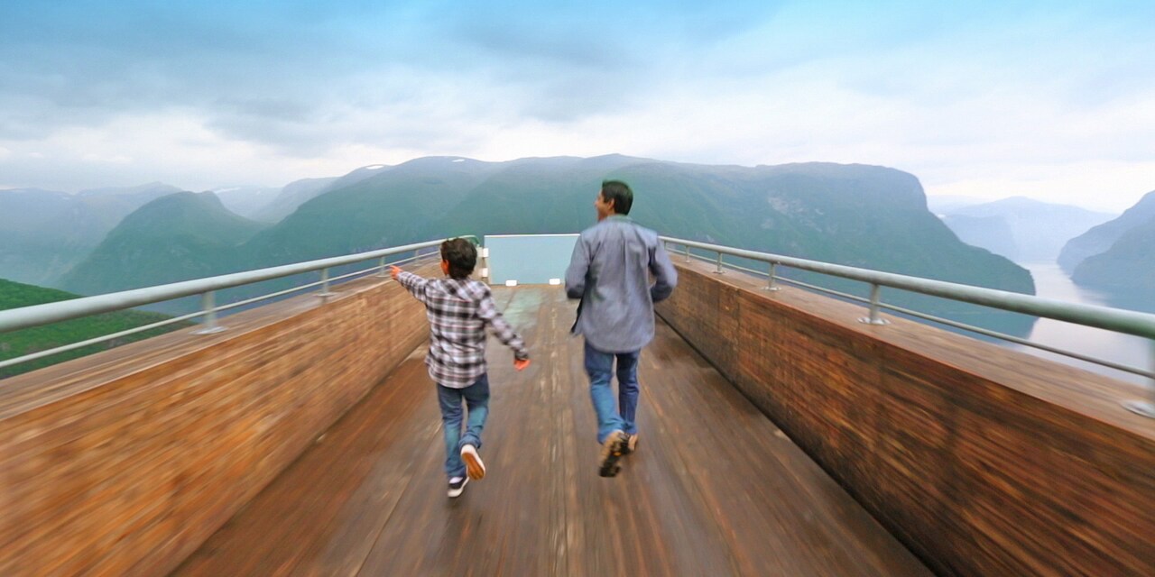 An excited father and son run down a wood-paneled skyway to view mountains and a winding river