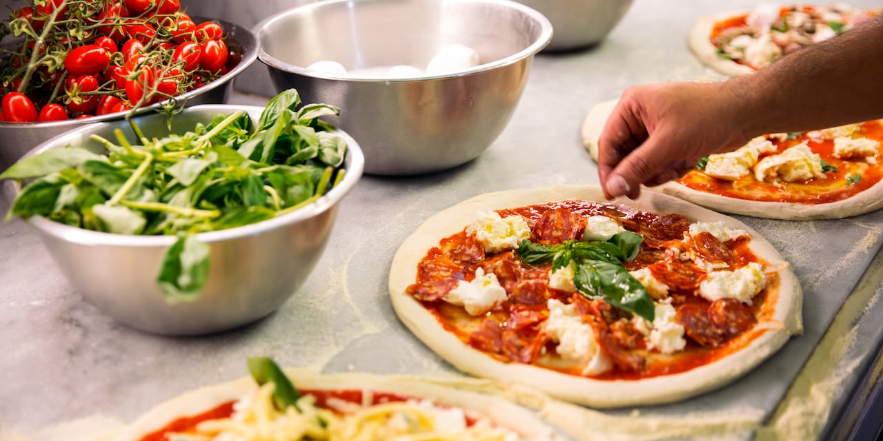 A hand sprinkles herbs on one of several pizzas lined up on a counter near metal mixing bowls filled with ingredients A hand sprinkles herbs on one of several pizzas lined up on a counter near metal mixing bowls filled with ingredients